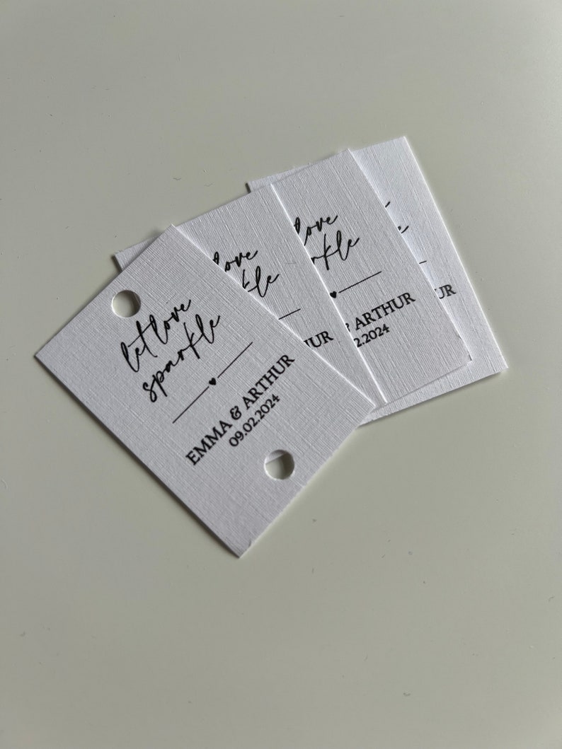 May include: White card stock tags with the text "Let love sparkle" and "Love like" in black script font. The tags also include the names "Emma & Arthur" and the date "09.02.2024" in black print.