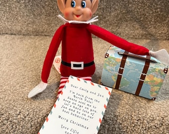 May include: A plush elf doll wearing a red suit with a white collar, a black belt, and red and white striped legs. The elf holds a small suitcase with a world map print and a white note with black text addressed to Emily and Jax from Elfie.