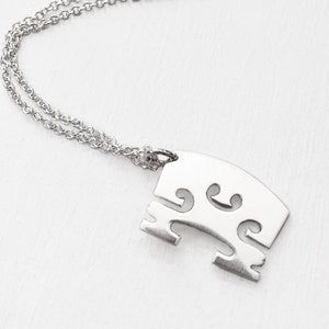 May include: A silver-colored necklace featuring a pendant shaped like a violin bridge. The pendant has a curved top with three decorative cutouts and two feet. The chain is a delicate, linked design.
