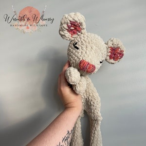 Puede incluir: Un juguete de peluche de ratón tejido a crochet con un cuerpo de color crema y orejas rosas y rojas. El ratón está sostenido por una mano con un tatuaje que dice "Better Together" con un diseño floral.
