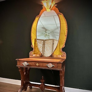 May include: A wooden vanity table featuring a large oval mirror with amber-tinted glass panels, a single drawer with decorative metal pulls, and a central finial. A stained glass light fixture is mounted at the top of the mirror frame.