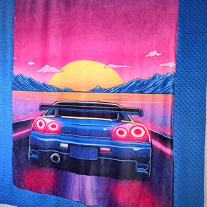 May include: A blue car blanket featuring a vibrant sunset scene with a car driving on a road. The blanket has a pink and purple sky, a yellow sun, and blue mountains. The car's taillights are glowing red.