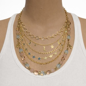 Puede incluir: Un collar dorado de varias capas con abalorios celestiales y cuentas rosas y azules. El collar presenta varios estilos de cadena, incluyendo delicados hilos con colgantes de estrella y luna. Las cuentas están intercaladas en el diseño.