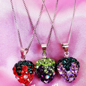 May include: Three silver chain necklaces with fruit-shaped pendants. The pendants are encrusted with colorful rhinestones, resembling a strawberry, blackberry, and grape. The background is a pink satin fabric.