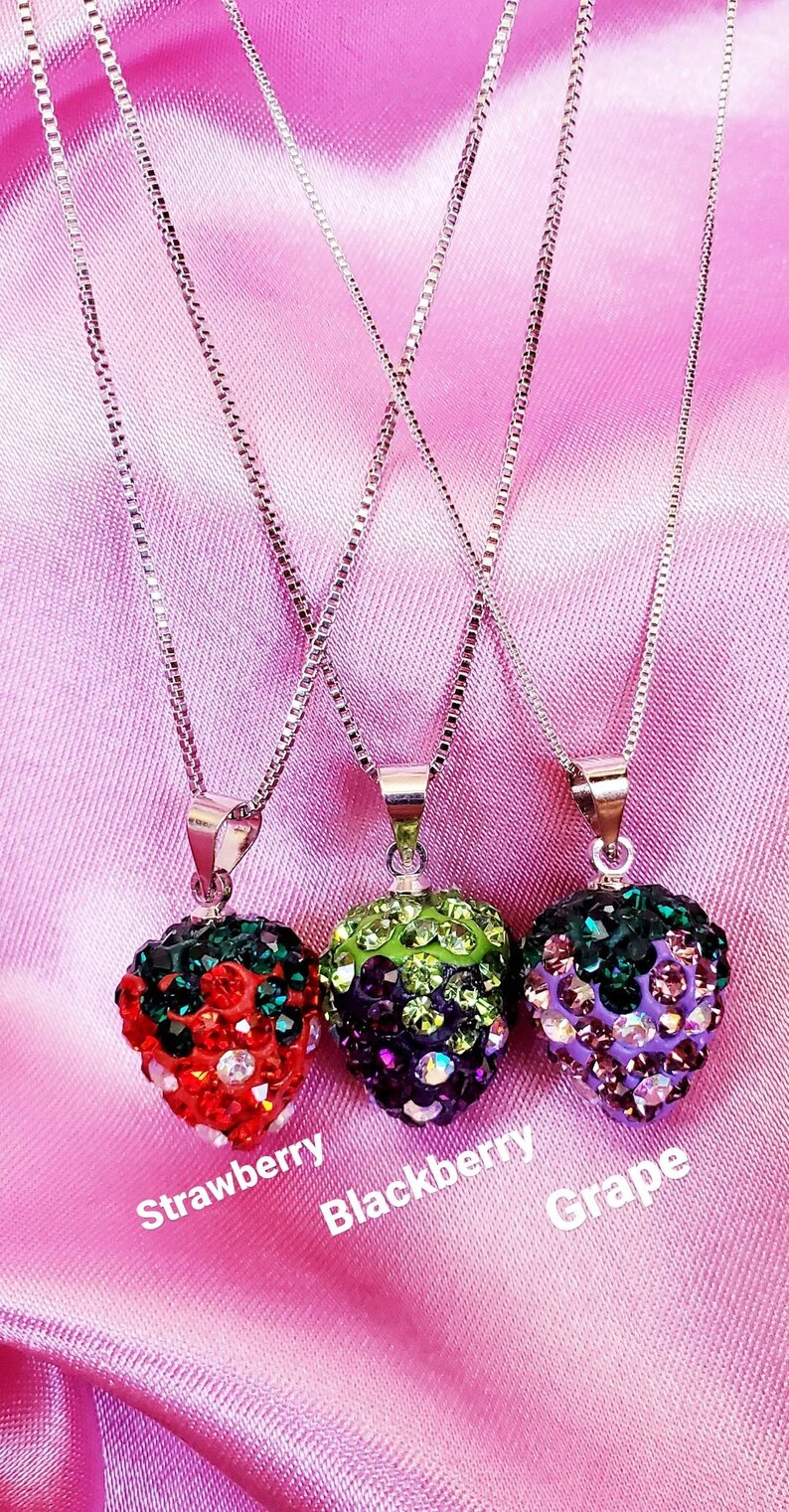 May include: Three silver chain necklaces with fruit-shaped pendants. The pendants are encrusted with colorful rhinestones, resembling a strawberry, blackberry, and grape. The background is a pink satin fabric.