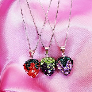 May include: Three silver chain necklaces with fruit-shaped pendants. The pendants are encrusted with colorful rhinestones, including a red strawberry, a green and purple berry, and a purple berry. The necklaces rest on a pink satin surface.