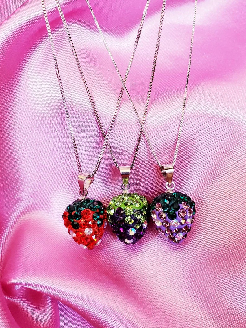 May include: Three silver chain necklaces with fruit-shaped pendants. The pendants are encrusted with colorful rhinestones, including a red strawberry, a green and purple berry, and a purple berry. The necklaces rest on a pink satin surface.