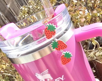 May include: A pink Stanley travel mug with a clear lid and a straw. The mug has a white logo that says "STANLEY". There are three strawberry-shaped charms hanging from the lid.
