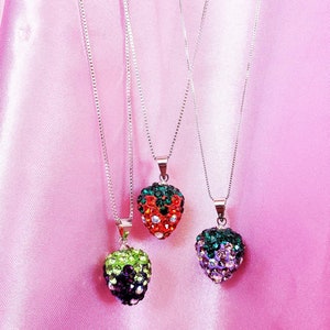 May include: Three strawberry-shaped pendant necklaces with silver chains. The pendants are encrusted with colorful rhinestones in red, green, and purple hues. The necklaces hang against a pink satin background.