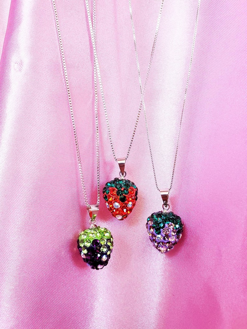 May include: Three strawberry-shaped pendant necklaces with silver chains. The pendants are encrusted with colorful rhinestones in red, green, and purple hues. The necklaces hang against a pink satin background.