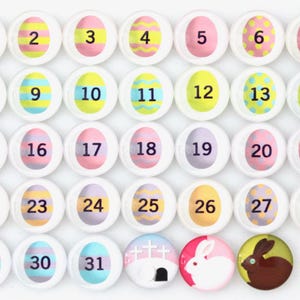 May include: A set of 31 round, white, Easter egg-shaped magnets with numbers 1 through 31. The magnets have different pastel colors and designs, including stripes, polka dots, and solid colors. The last magnet has a picture of a white bunny rabbit.