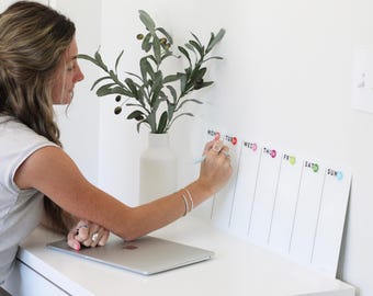 May include: A white dry-erase board with a weekly calendar layout. The board has the days of the week labeled: Mon, Tues, Wed, Thurs, Fri, Sat, Sun. Each day has a colored circle next to it. A person is writing on the board with a pen.