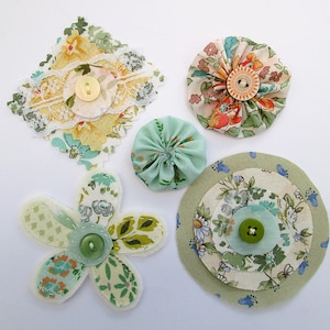 May include: A selection of handmade fabric flowers and embellishments. The items feature floral patterns in green, yellow, and blue, with button centres. Some have lace trim. Measurements are in centimetres.