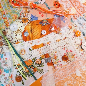 May include: A selection of craft materials featuring patterned fabric pieces, lace, buttons, beads, and ribbons. The colour scheme is predominantly orange and cream, with floral designs. A small charm is visible, inscribed with the word "Dream".