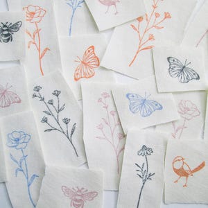 May include: A selection of small, off-white fabric squares featuring nature-themed illustrations. Designs include bees, butterflies, flowers, and birds in black, orange, pink, and blue. The fabric squares are arranged across a surface.