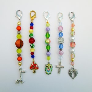 May include: Five beaded charms with silver-tone lobster clasps. The charms feature colourful beads in various shapes and sizes. Each charm has a unique pendant: a giraffe, a mushroom, an owl, a cross, and a pair of wings.