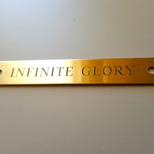 May include: A rectangular, gold-colored metal plaque with rounded corners and two holes. The words "INFINITE GLORY" are engraved in the center. The plaque is set against a white background.