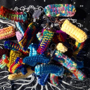 May include: A collection of colorful, handmade crochet keychains. The keychains feature a variety of vibrant colors, including blue, yellow, red, and green. Each keychain has a metal clasp for easy attachment. The keychains are displayed on a black background.