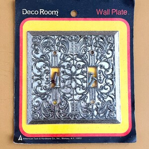 May include: A decorative silver-colored double light switch wall plate with an ornate floral design. The packaging reads "Deco Room" and "Wall Plate." The packaging is black, yellow, and red. The manufacturer's name and address are printed at the bottom.