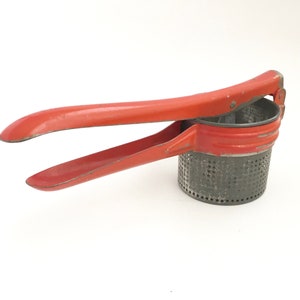 May include: A vintage red and gray potato ricer. The kitchen utensil has a perforated metal cup and long, red handles for pressing potatoes. The ricer is designed to create smooth mashed potatoes. The handles are approximately 10 inches long.