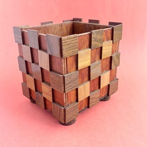 May include: A square wooden container with a woven pattern of various shades of brown. The container sits on four small, round feet. The interior of the container is visible, showing a lighter wood tone. The background is a solid pink color.