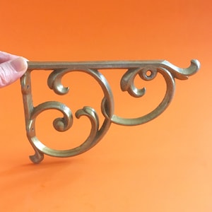 May include: A brass-colored decorative shelf bracket with an ornate, scrolled design. The bracket has a flat top and a curved, flowing pattern, with a small hole for mounting. The bracket is held against an orange background.