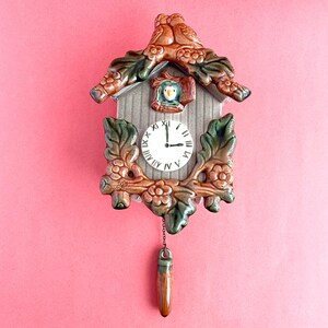 May include: A ceramic cuckoo clock with a pink background. The clock features a white clock face with black Roman numerals and black hands. The clock is decorated with brown, green, and tan floral and bird details.