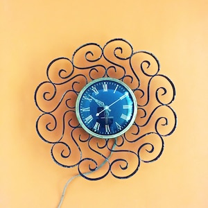 May include: A vintage wall clock with a dark blue face and Roman numeral hour markers. The clock is encased in a gold-colored metal frame and surrounded by a decorative, black wrought iron scrollwork design. A cord extends from the clock.