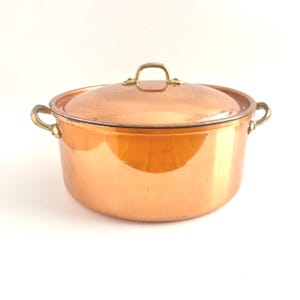 May include: A round copper pot with a matching lid, featuring two brass side handles and a brass handle on the lid. The exterior surface is polished copper with a warm metallic sheen, while the handles are secured with visible metal rivets.