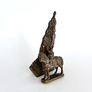 May include: A small bronze sculpture of a rider on horseback, holding a miniature church-like structure above their head. The sculpture is detailed and intricate, with a textured surface.