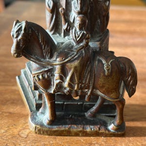 May include: A bronze sculpture of a woman riding a horse. The woman is wearing a long robe and has her hand raised in the air. The horse is standing on a base with steps.