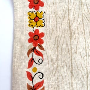 May include: Close-up of a cream-colored fabric with a vertical floral border. The border features red and yellow flowers with brown centers, along with brown leaves and stems. The background has a subtle, textured pattern.