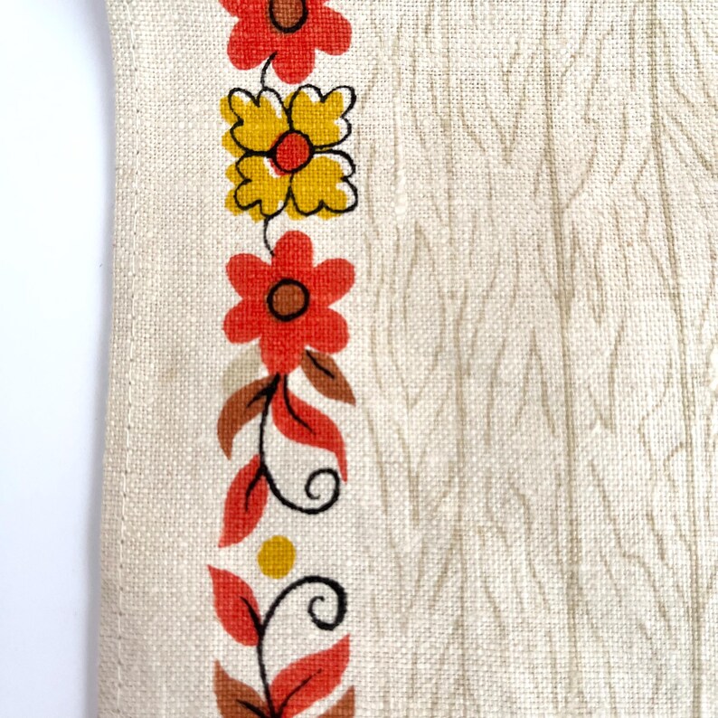 May include: Close-up of a cream-colored fabric with a vertical floral border. The border features red and yellow flowers with brown centers, along with brown leaves and stems. The background has a subtle, textured pattern.
