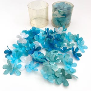 May include: A collection of blue and white fabric flower embellishments. The flowers are in various shades of blue, with some white petals. They are scattered on a white surface, with two clear plastic containers in the background.