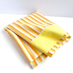 May include: A folded yellow and orange striped fabric with a solid yellow border. The fabric is folded in half and the yellow border is visible on the outside.