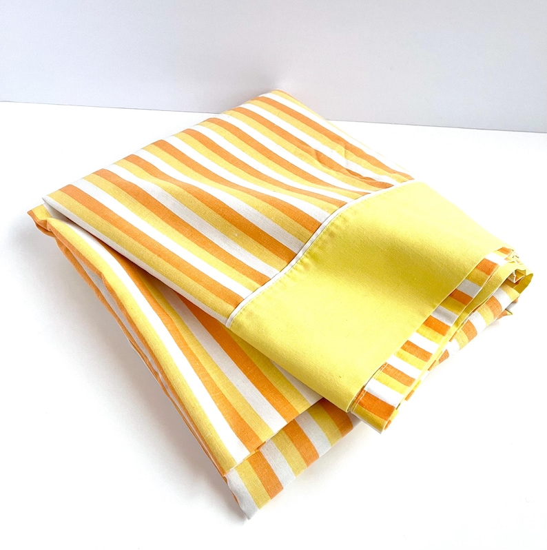 May include: A folded yellow and orange striped fabric with a solid yellow border. The fabric is folded in half and the yellow border is visible on the outside.