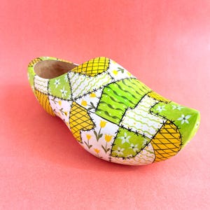 May include: A wooden clog featuring a patchwork design of yellow, white, and green squares. The pattern includes yellow tulips, white flowers, wavy green lines, and yellow cross-hatching, all outlined with black stitches on a light background.