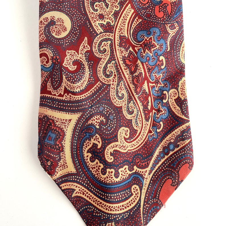 May include: A red, blue, and beige paisley patterned tie. The tie has a pointed tip and a classic design.