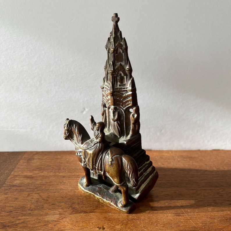 May include: A bronze sculpture of a person on horseback, with a tall, ornate building behind them. The sculpture is detailed and intricate, with a realistic depiction of the horse and rider.
