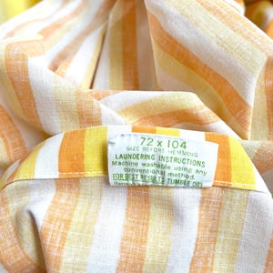 May include: A white fabric with orange and yellow stripes. The fabric has a label that reads "72 x 104 SIZE BEFORE HEMMING LAUNDERING INSTRUCTIONS Machine washable using any conventional method. FOR BEST RESULTS TUMBLE DRY Reme"