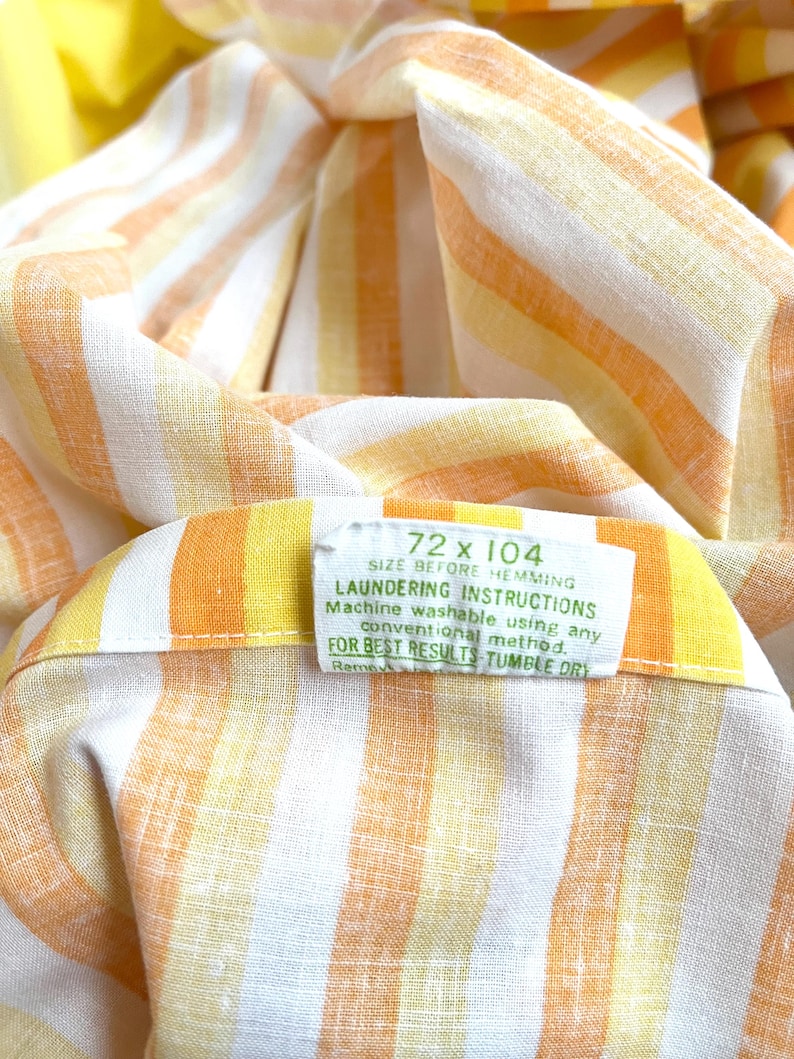 May include: A white fabric with orange and yellow stripes. The fabric has a label that reads "72 x 104 SIZE BEFORE HEMMING LAUNDERING INSTRUCTIONS Machine washable using any conventional method. FOR BEST RESULTS TUMBLE DRY Reme"