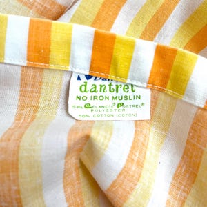 May include: A white fabric with orange and yellow stripes. The fabric has a label that reads "dantret NO IRON MUSLIN 50% ELANESE PORTREL POLYESTER 50% COTTON (COTON)"