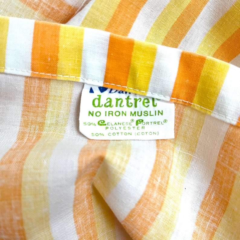 May include: A white fabric with orange and yellow stripes. The fabric has a label that reads "dantret NO IRON MUSLIN 50% ELANESE PORTREL POLYESTER 50% COTTON (COTON)"
