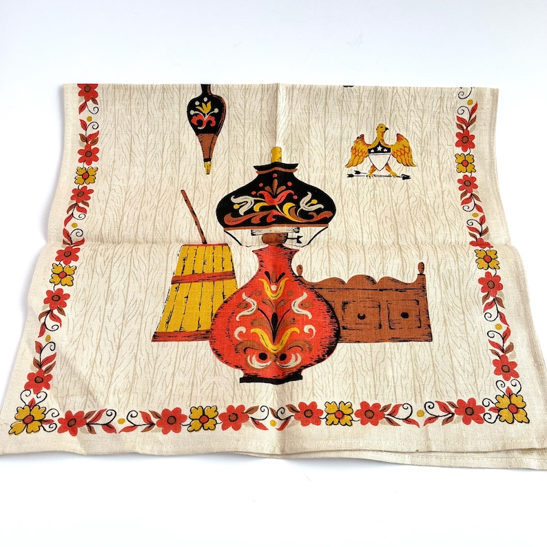 May include: Cream-colored vintage kitchen towel with a decorative border of red and yellow flowers. The center features a lamp, a broom, and a cabinet in shades of orange, black, and brown. An eagle emblem is also present.