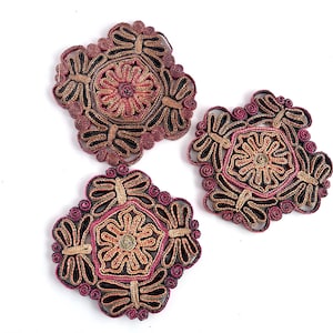 May include: Three decorative coasters with intricate floral designs. Each coaster features a central flower pattern in shades of pink, tan, and gold, surrounded by a dark brown and burgundy border. The coasters are arranged on a white surface.