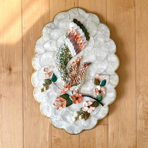 May include: A white oval-shaped wall decor with a raised design of a bird with a long tail made of seashells. The bird is surrounded by flowers and leaves, also made of seashells.