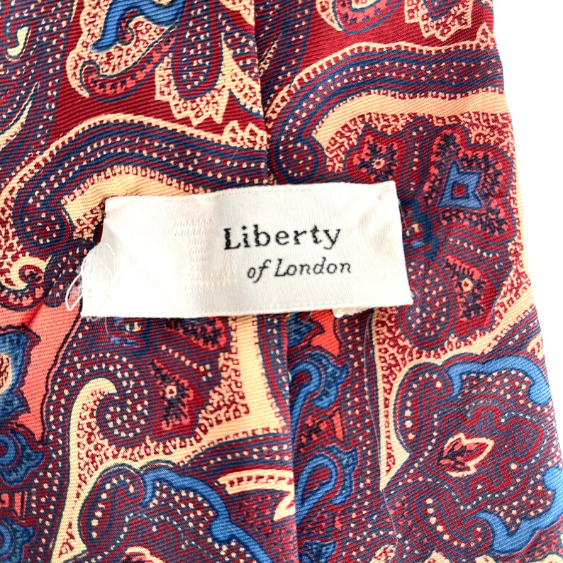 May include: A red, blue, and cream paisley patterned tie with a white tag that reads "Liberty of London".