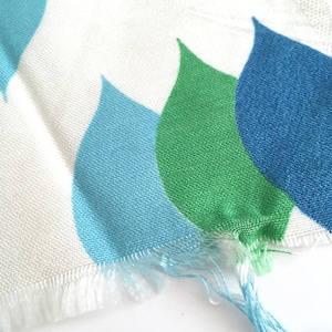 May include: Close-up of a white fabric with a blue, green, and light blue teardrop pattern. The fabric has a frayed edge.