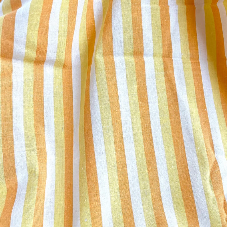 May include: White and yellow striped fabric with a subtle texture. The fabric is folded and draped, showing the stripes in a diagonal pattern.