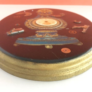 May include: A round, dark red, wooden plaque with a gold trim. The plaque features a vintage clock face with gold accents and a blue base. The clock is surrounded by various small, metallic gears and other clock parts.